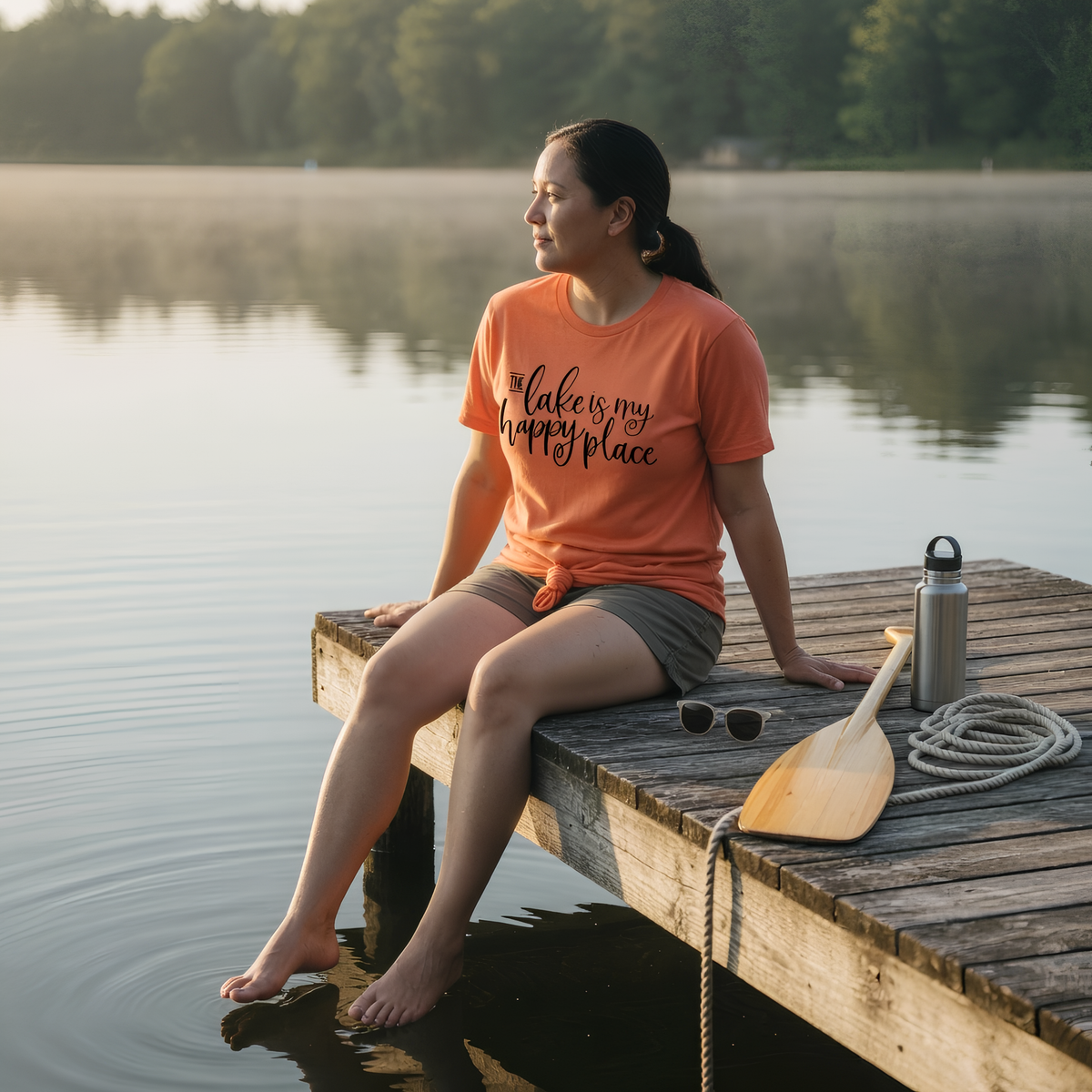 A woman in an orange t-shirt sits on a wooden dock, with a paddle and water bottle nearby, overlooking a calm lake surrounded by trees.