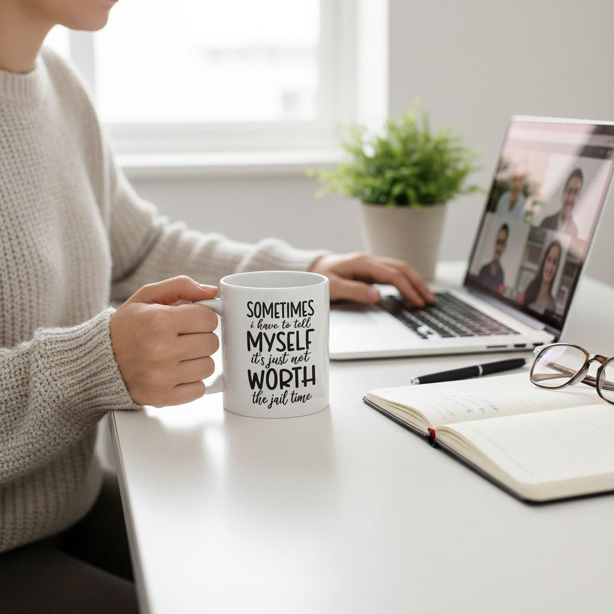 A person is sitting at a desk, holding a mug with the text "Sometimes I just feel worth the jail time" printed on it. The desk also has a laptop, a notebook, and a pair of glasses.