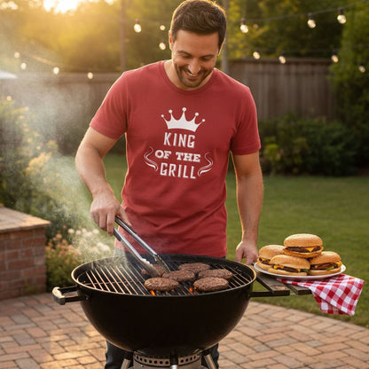 A man in a red t-shirt is grilling hamburgers on a barbecue grill, surrounded by a patio setting with string lights and greenery.