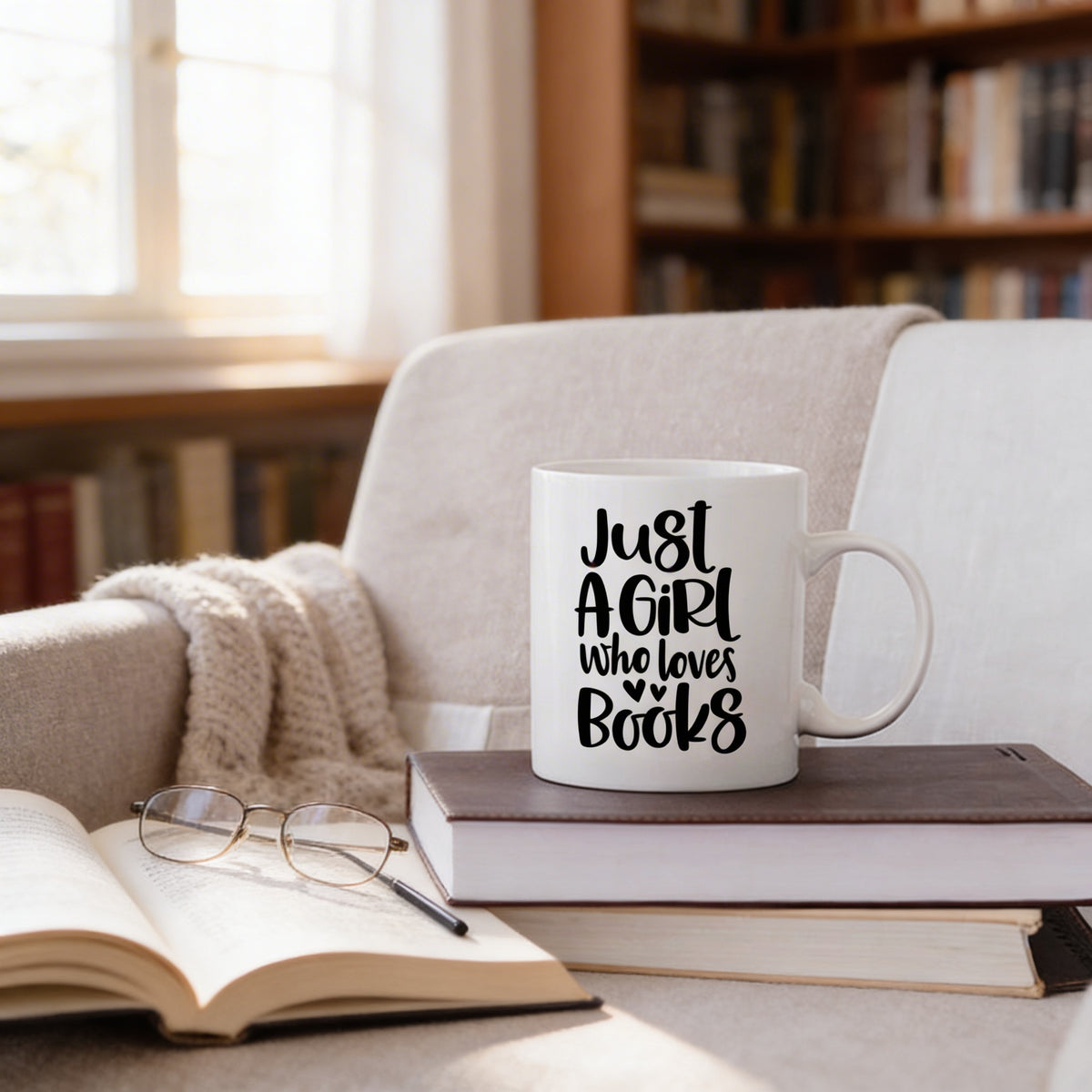 A white mug with the text "Just a girl who loves books" sits on top of a stack of books, with a pair of glasses and a cozy blanket nearby.