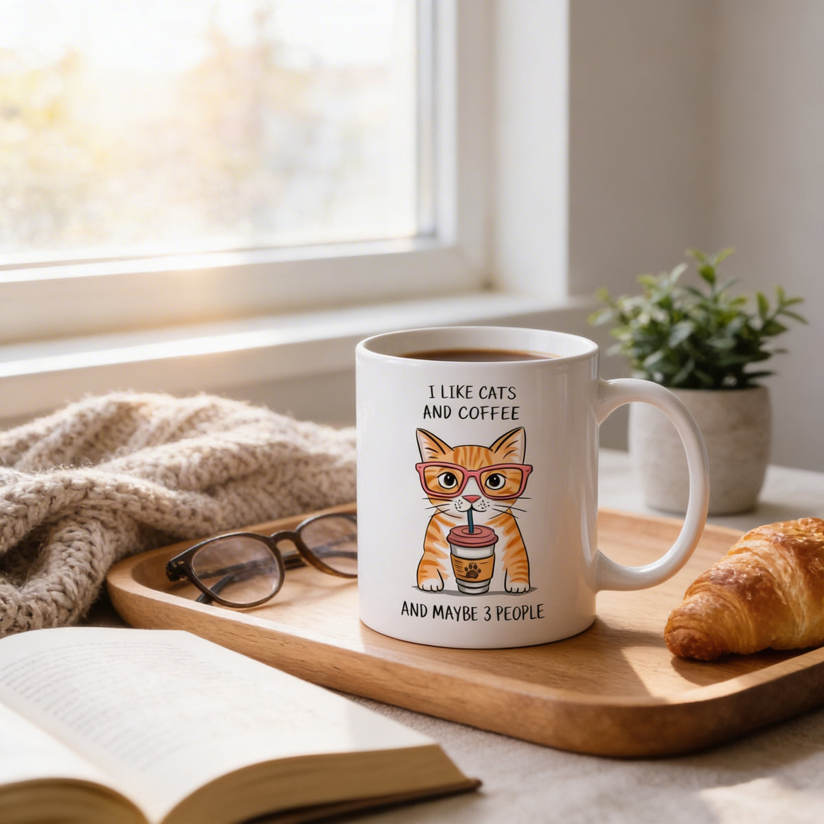 a mug with a cat wearing glasses and holding a coffee cup, sitting on a wooden tray next to a book and a croissant.