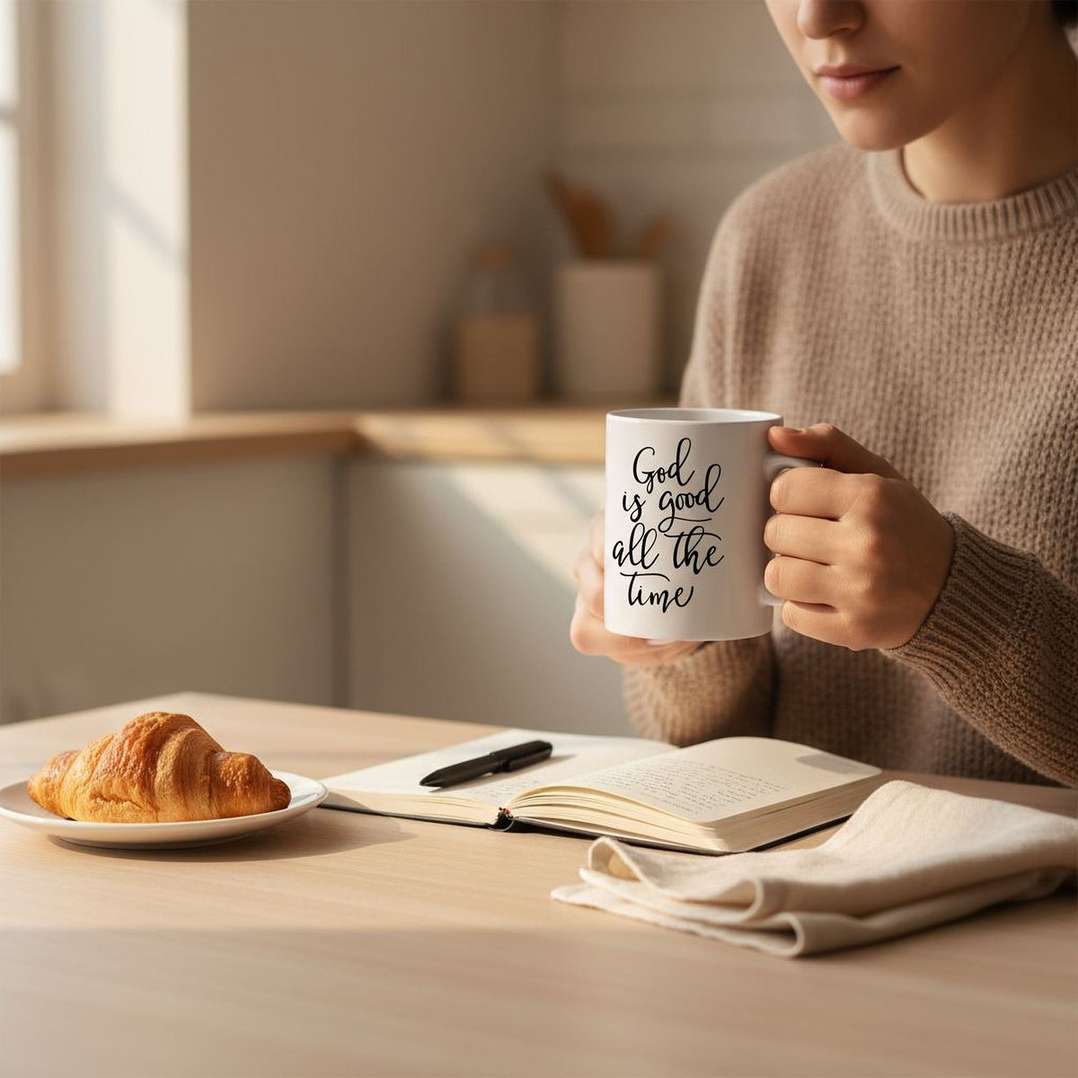 A person is sitting at a table, holding a mug with the text "God is good all the time" written on it. There is a plate of food, specifically a croissant, in front of the person.