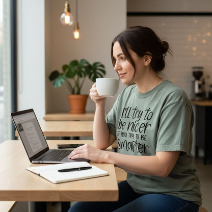 A woman wearing a green t-shirt with text is sitting at a table in a coffee shop, working on a laptop and holding a coffee mug.