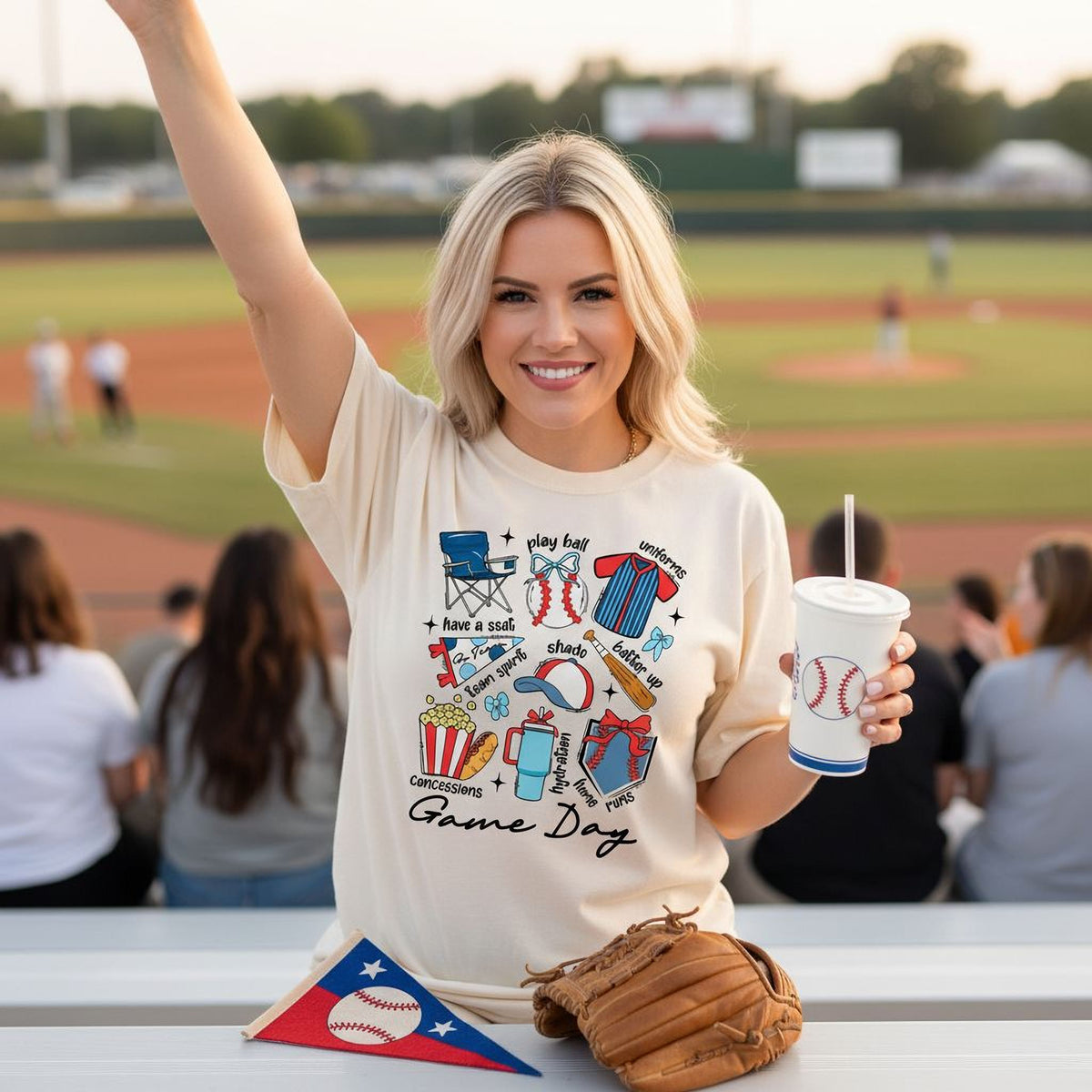 A smiling woman wearing a white t-shirt with a baseball-themed design is holding a cup and a baseball glove, standing in front of a baseball field with spectators in the background.