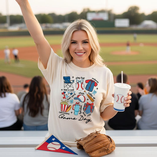 A smiling woman wearing a white t-shirt with a baseball-themed design is holding a cup and a baseball glove, standing in front of a baseball field with spectators in the background.