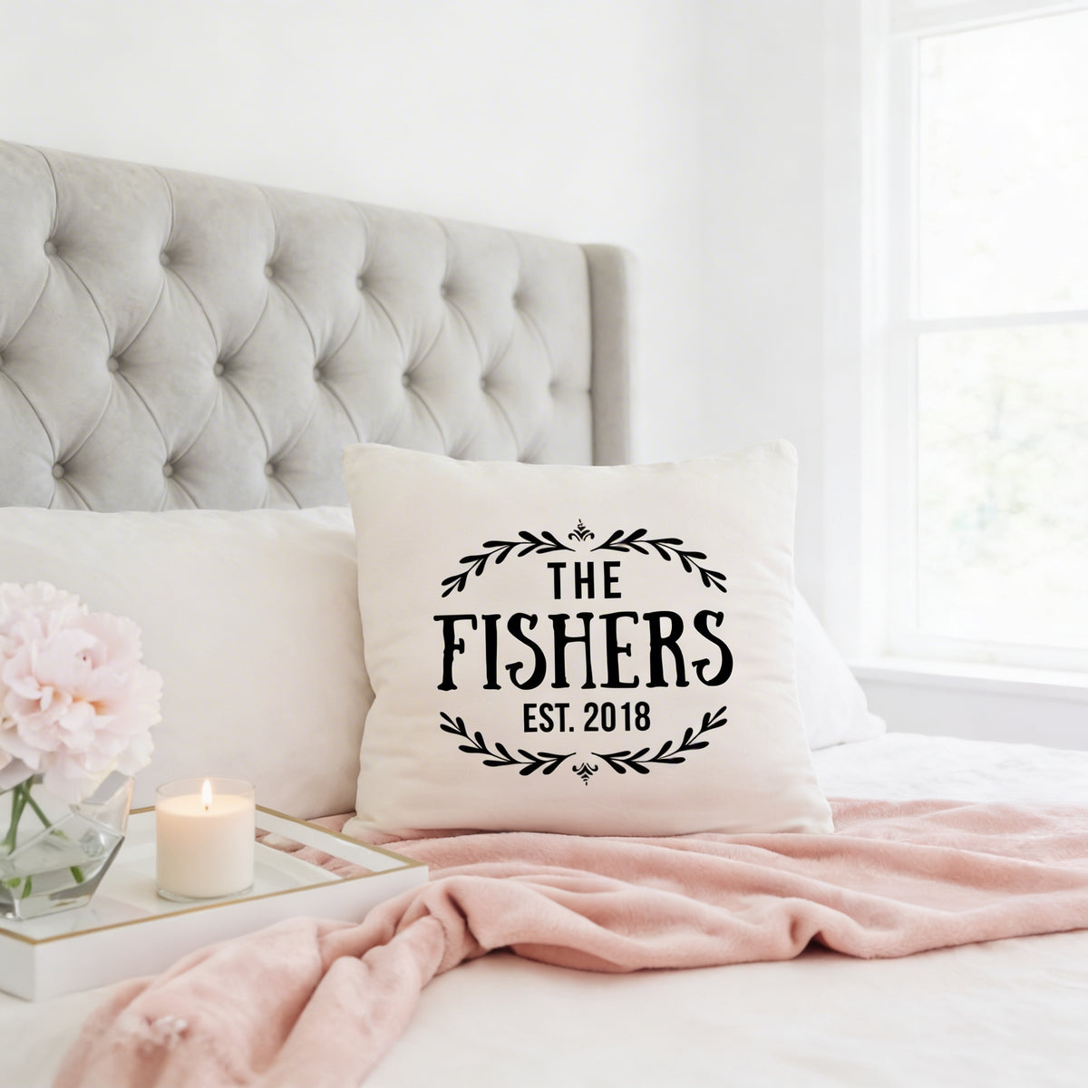 A white tufted headboard, a white pillow with the text "THE FISHER'S EST. 2018" printed on it, and a pink blanket on a bed.