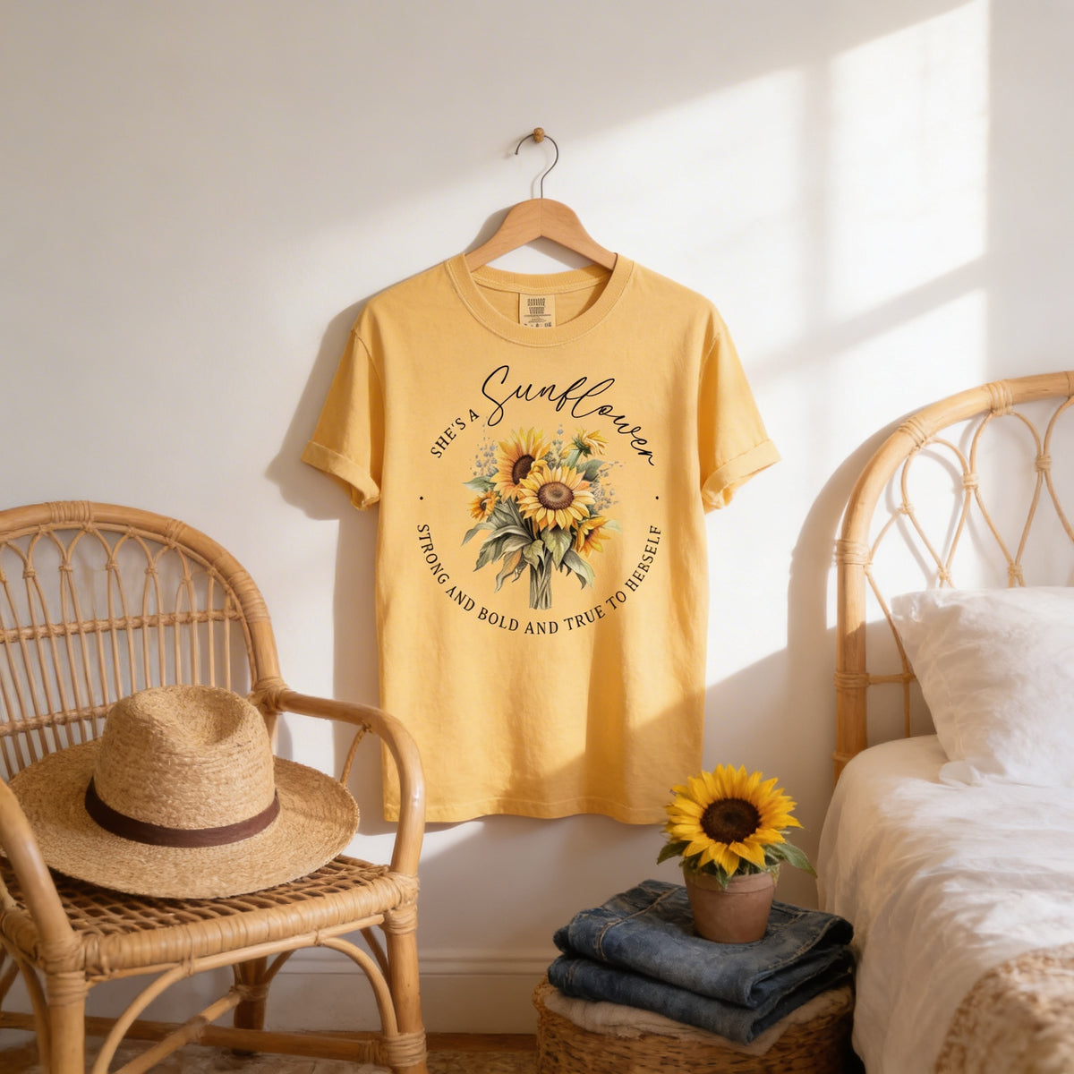 a yellow t-shirt with a sunflower design hanging on a wooden hanger, accompanied by a straw hat and a small potted sunflower on a bedside table.