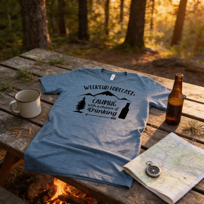A t-shirt with a camping-themed design, a mug, a bottle, a compass, and a map are arranged on a wooden table, with a campfire visible in the background.