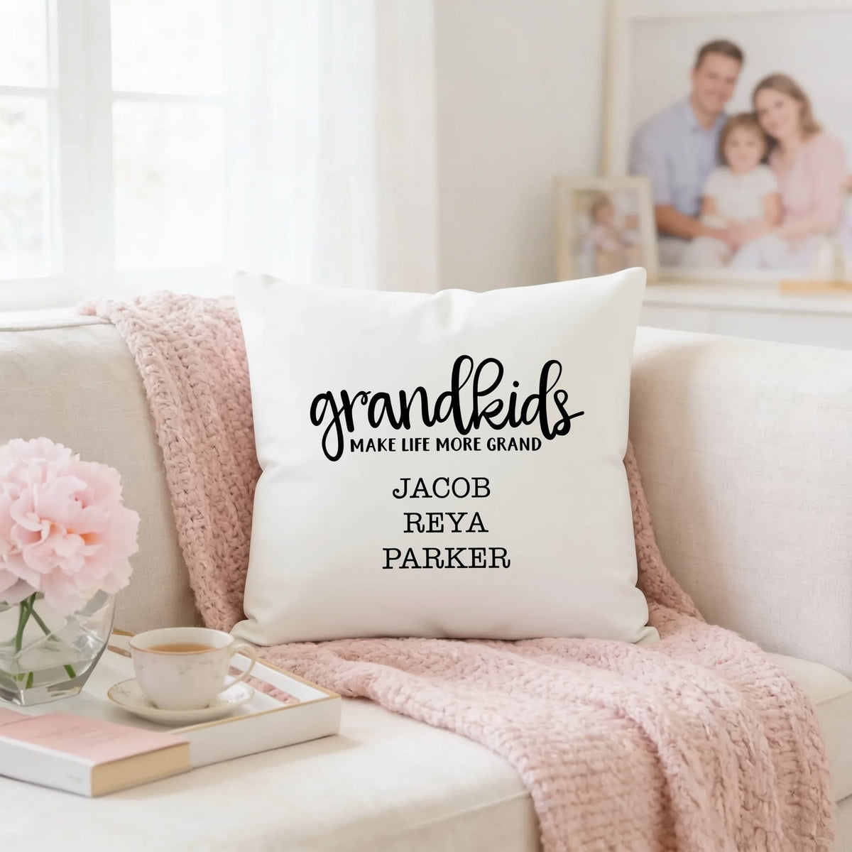 A white throw pillow with the text "Grandkids make life more grand" printed on it, sitting on a couch with a pink blanket and a cup of tea nearby.