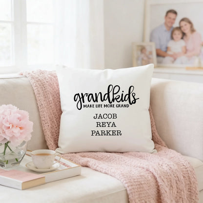 A white throw pillow with the text "Grandkids make life more grand" printed on it, sitting on a couch with a pink blanket and a cup of tea nearby.