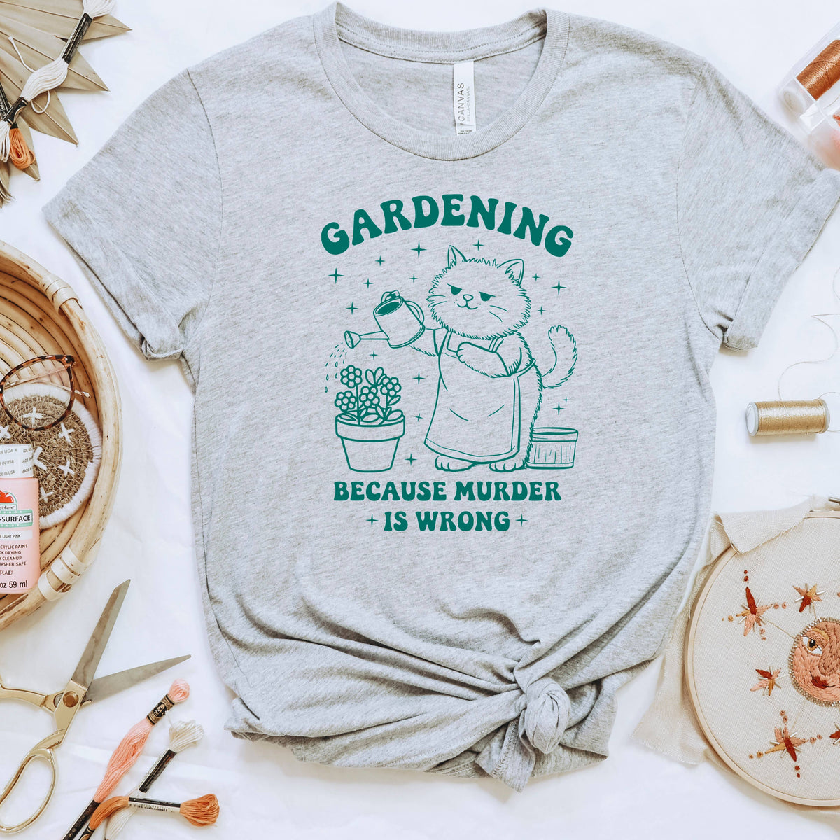 a grey t-shirt with a graphic design depicting a cat watering a plant, accompanied by the text "GARDENING BECAUSE MURDER IS WRONG".