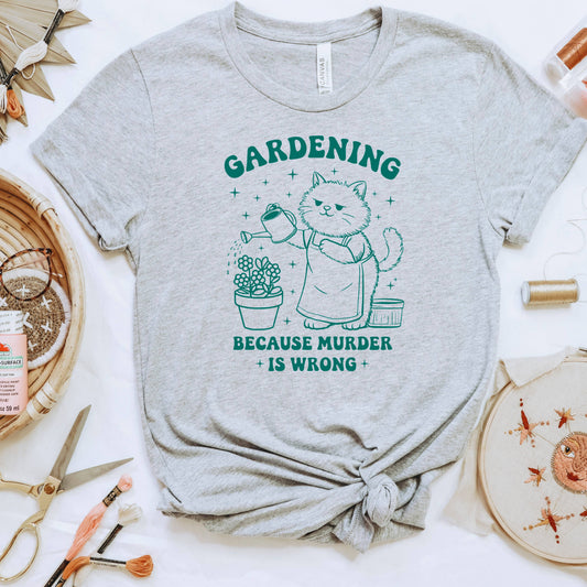 a grey t-shirt with a graphic design depicting a cat watering a plant, accompanied by the text "GARDENING BECAUSE MURDER IS WRONG".