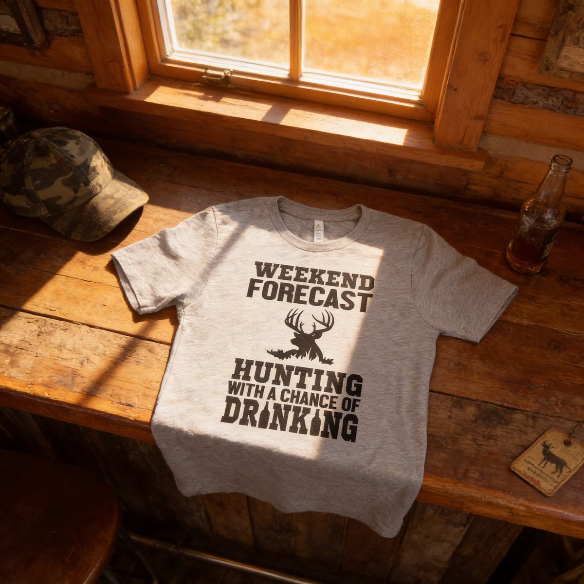 A gray t-shirt with a humorous text printed on it, placed on a wooden surface next to a bottle and a hat.