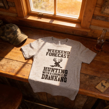 A gray t-shirt with a humorous text printed on it, placed on a wooden surface next to a bottle and a hat.