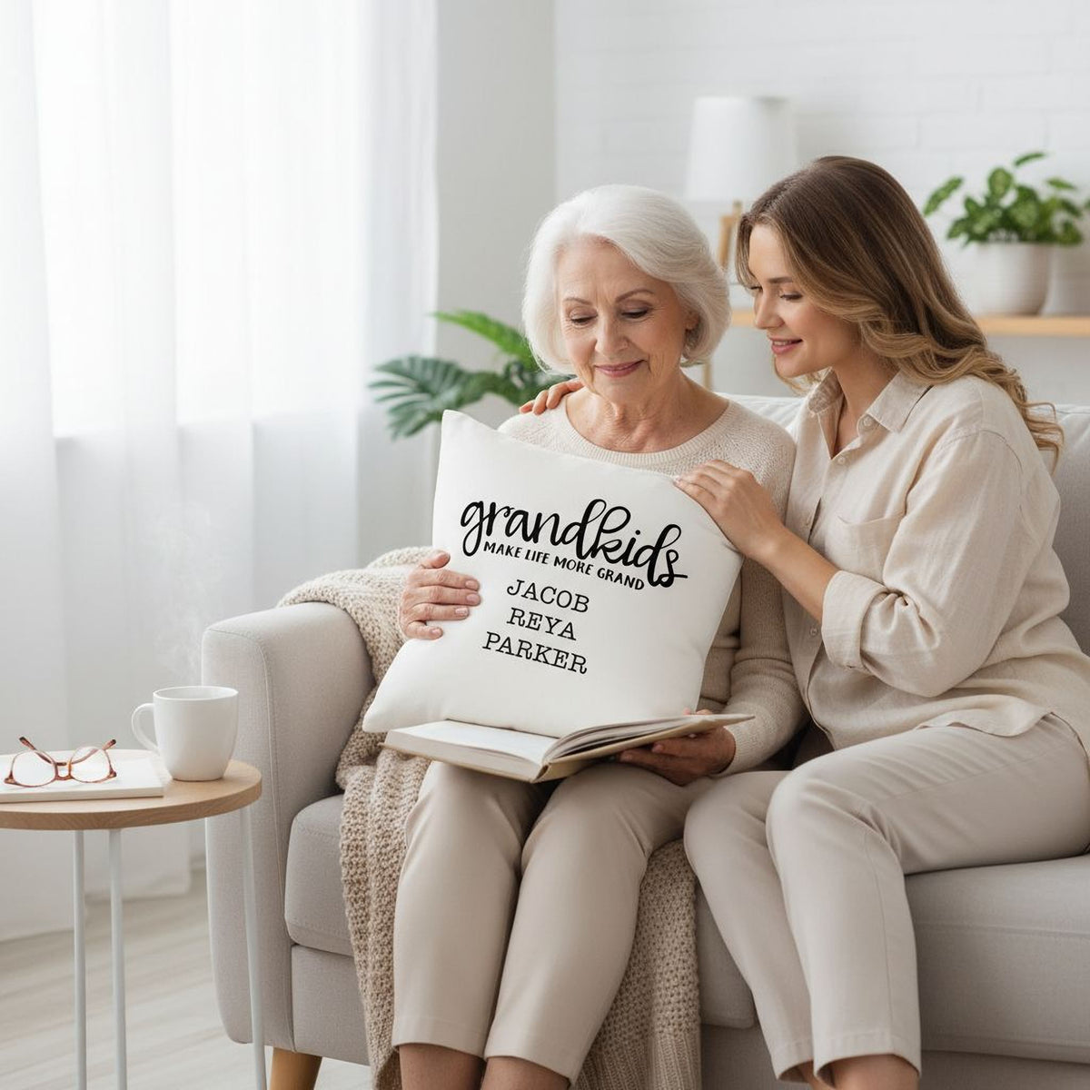 Two women sitting on a couch, one older and one younger, are looking at a book together. The older woman is holding a pillow with the text "Grandkids" written on it.