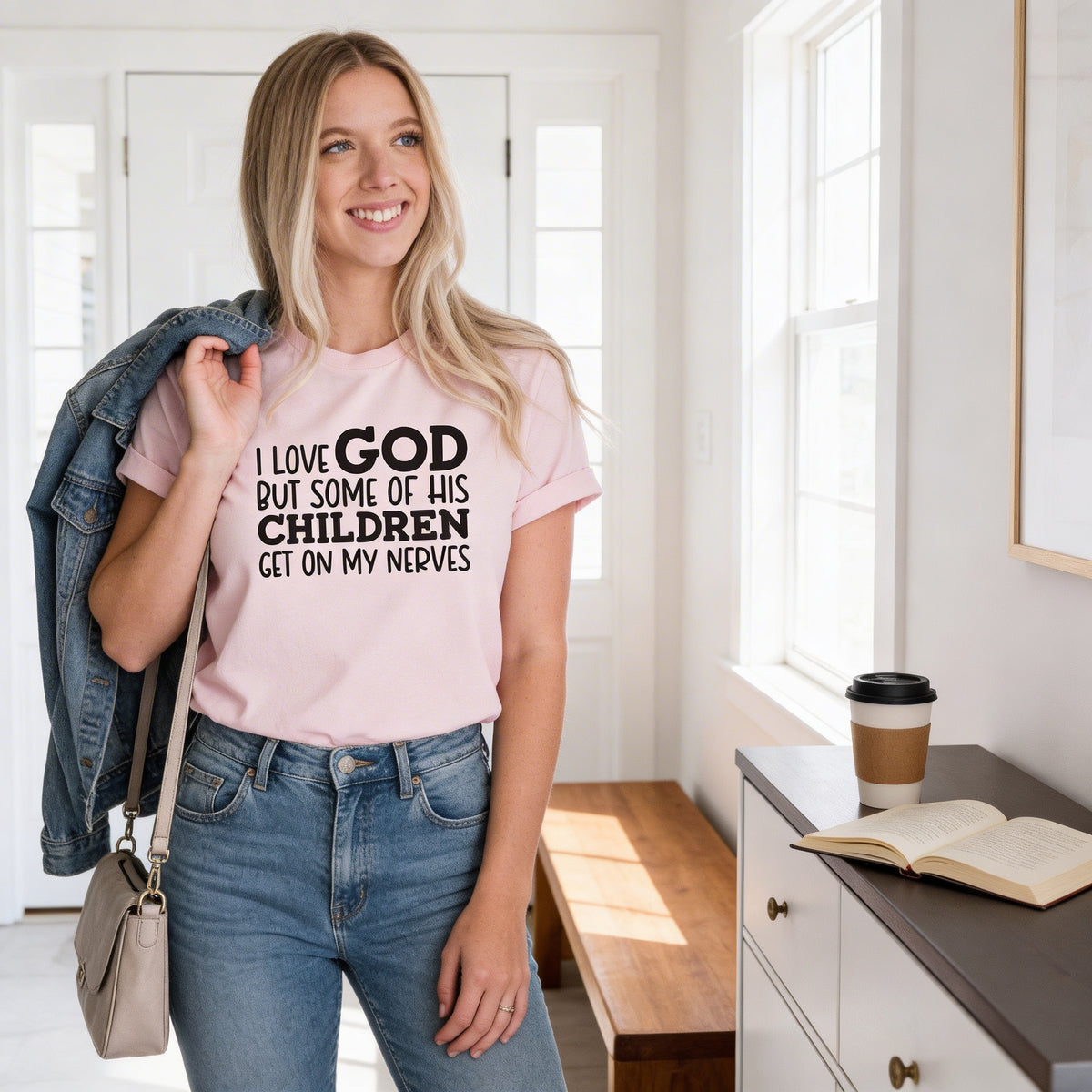 A woman wearing a pink t-shirt with text stands in a room, smiling and holding a denim jacket over her shoulder.