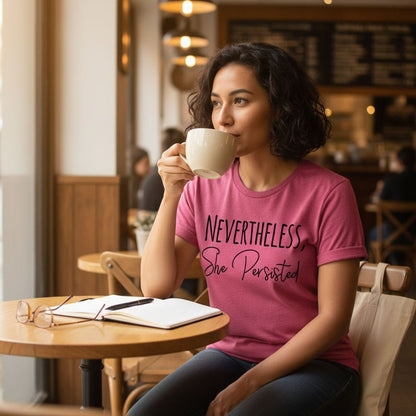 A woman wearing a pink t-shirt with the text "NEVERTHELESS She Persisted" is sitting at a table in a coffee shop, sipping a cup of coffee.
