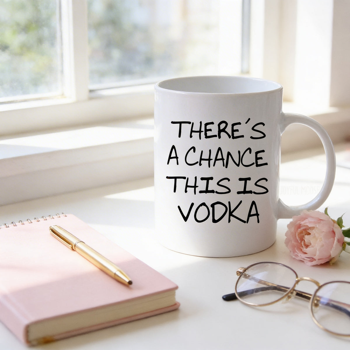 a white mug with the text "THERE'S A CHANCE THIS IS VODKA" printed on it, placed on a white surface next to a pink notebook, a gold pen, and a pair of glasses.