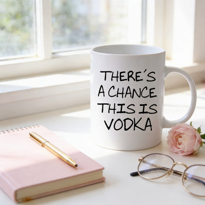 a white mug with the text "THERE'S A CHANCE THIS IS VODKA" printed on it, placed on a white surface next to a pink notebook, a gold pen, and a pair of glasses.