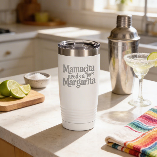 A white tumbler with the text "Mamacita needs a Margarita" printed on it, placed on a kitchen counter next to a cocktail shaker and a glass with a lime wedge.