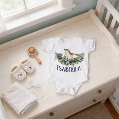 A baby's white onesie with a bird design, a pair of white booties, and a baby rattle are arranged on a white dresser.