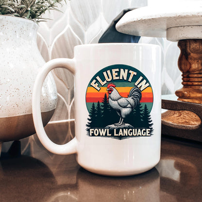 A white mug with a colorful graphic design featuring a rooster and the text "FLUENT IN FOWL LANGUAGE" on it.
