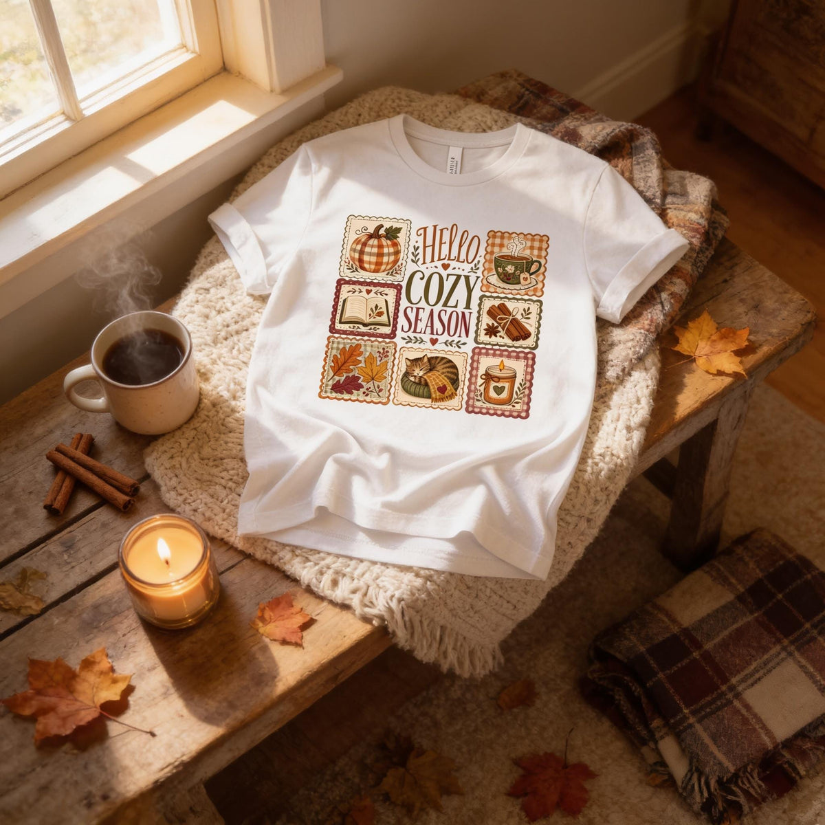 A cozy t-shirt with a graphic design, a mug of coffee, a candle, and a plaid blanket are arranged on a wooden surface, creating a warm and inviting atmosphere.
