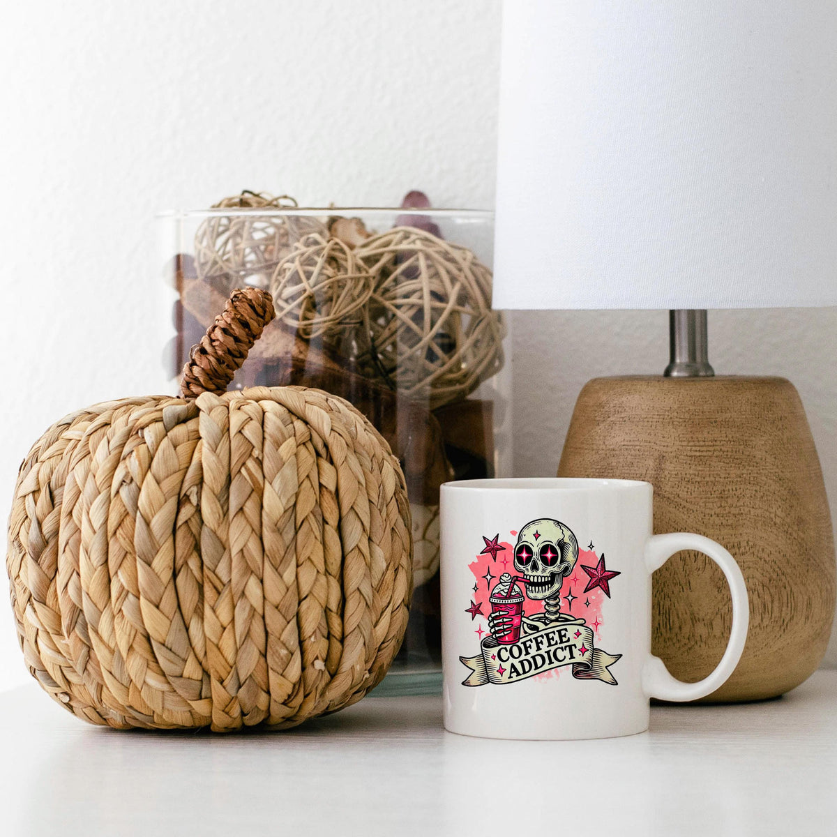 a white coffee mug with a skull design and the text "Coffee Addict" printed on it, placed on a white surface next to a woven pumpkin decoration and a lamp.