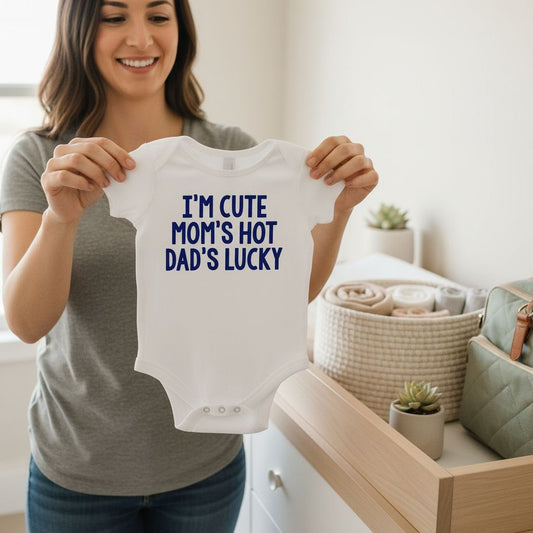 A woman is holding up a white onesie with the text "I'm cute mom's hot dad's lucky" printed on it.