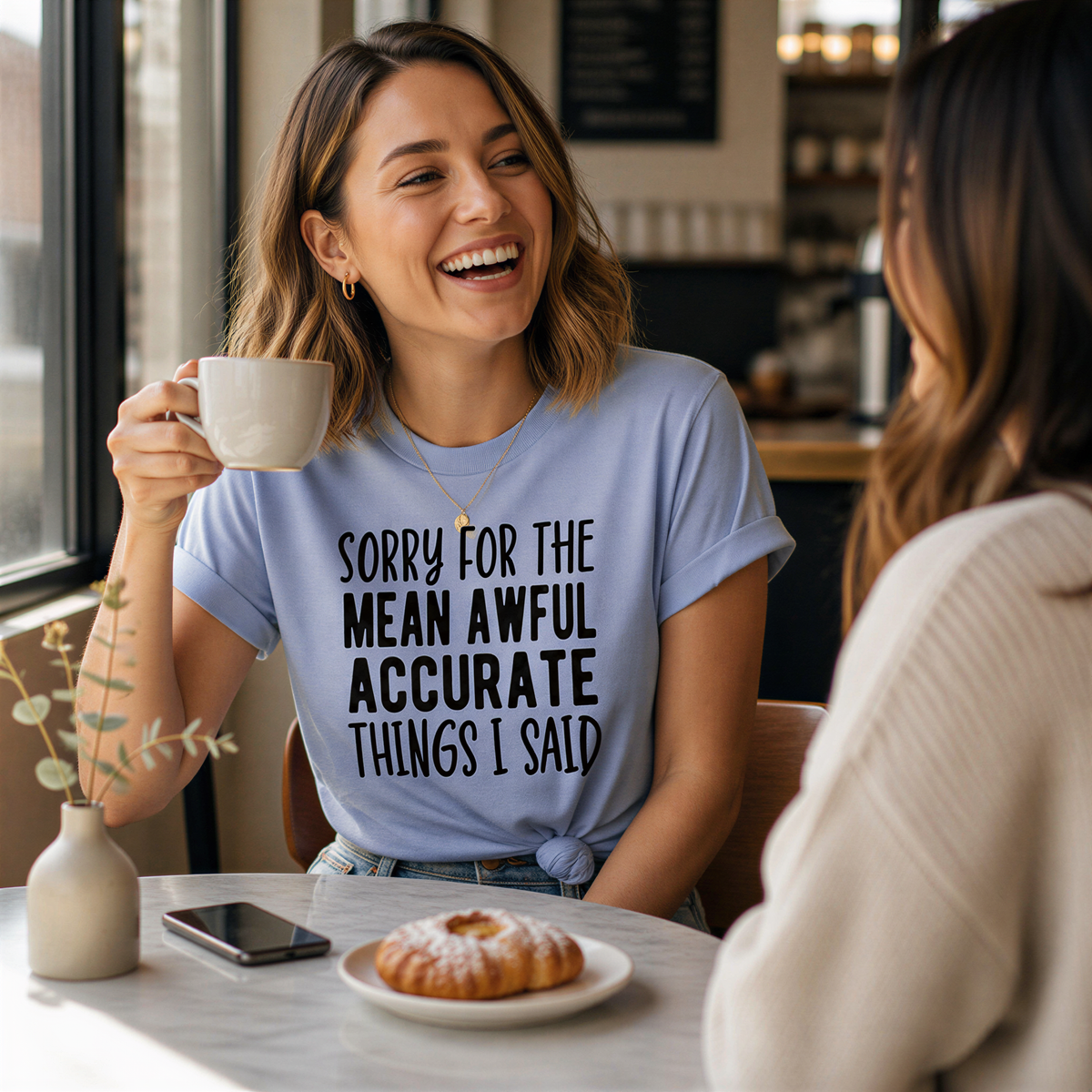 A woman wearing a blue t-shirt with text is laughing and smiling while holding a coffee mug, with another woman sitting across from her at a table with a pastry and a coffee cup.