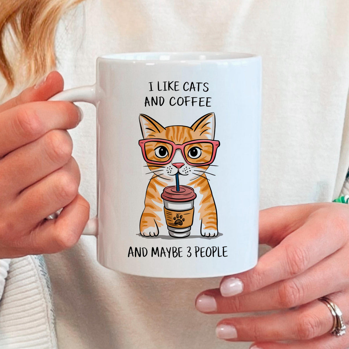 A person is holding a white mug with an illustration of a cat wearing glasses and holding a coffee cup.