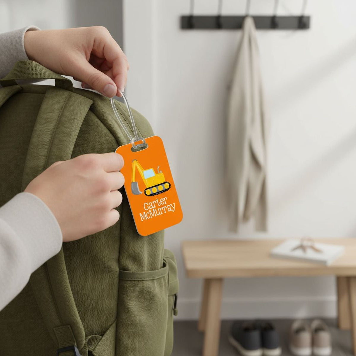 A person's hands holding a green backpack with an orange luggage tag attached, displaying the name "Carrie McMurray" and an illustration of a construction vehicle.