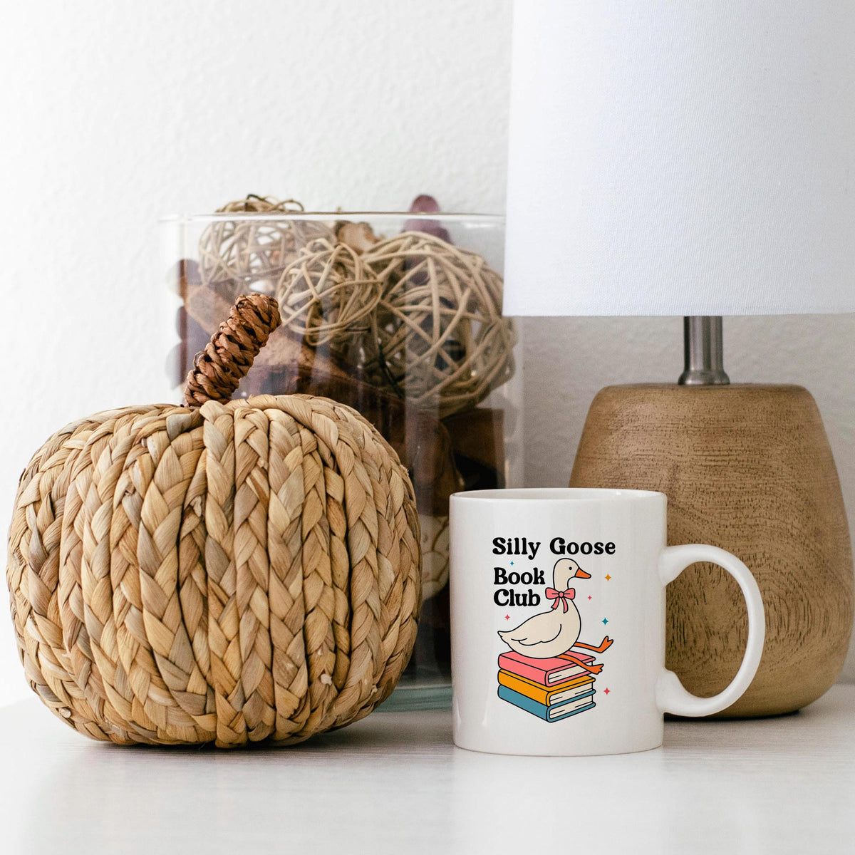 a white mug with a design of a duck sitting on a stack of books, placed next to a woven pumpkin decoration and a lamp.