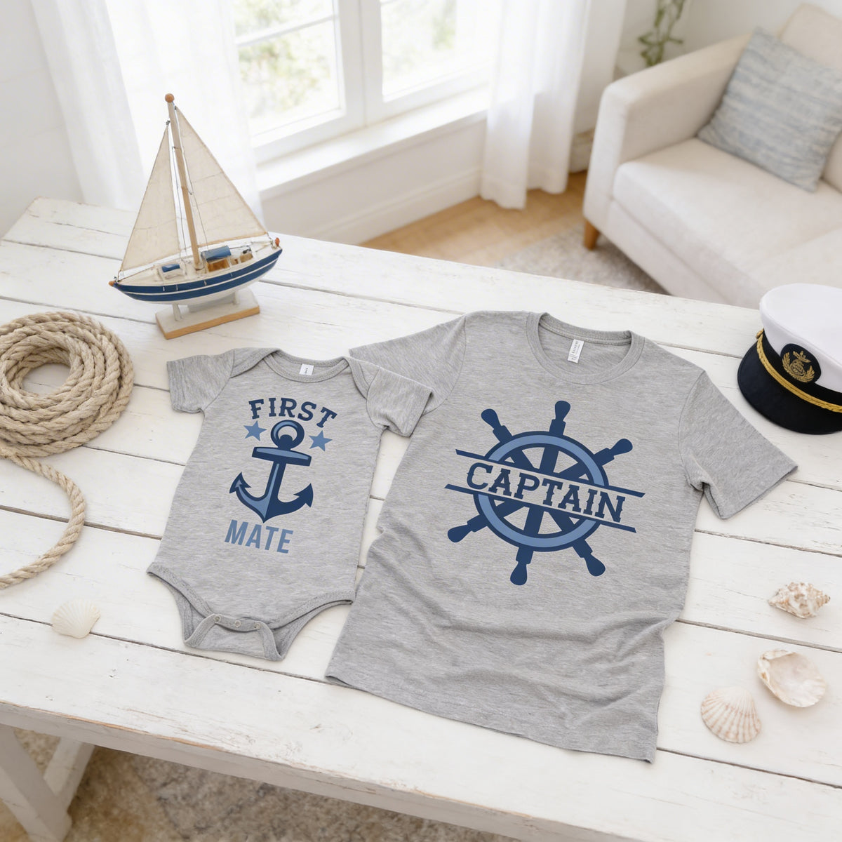 a gray t-shirt with a blue anchor and a smaller gray onesie with an anchor design, both labeled "FIRST MATE" and "CAPTAIN" respectively. The items are placed on a white wooden table, along with a small sailboat model, a hat, and some seashells.