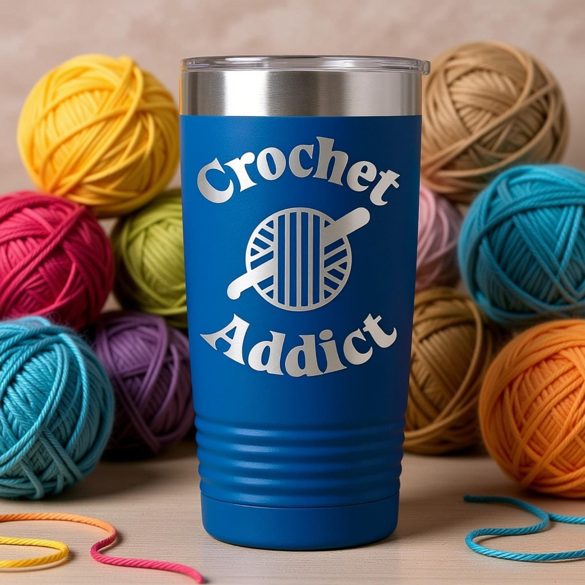 A blue insulated tumbler with the text "Crochet Addict" printed on it, surrounded by various colored yarn balls.