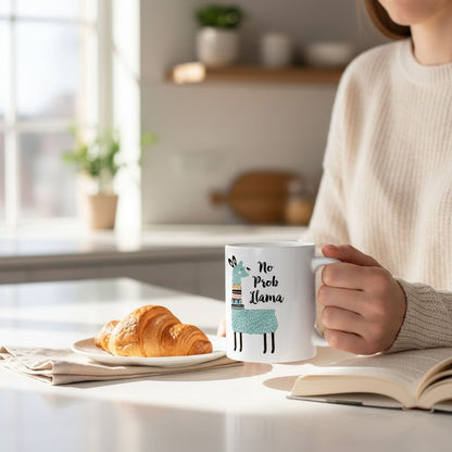 A person is sitting at a table, holding a mug with a cartoon llama design and the text "No Prob Llamas" printed on it. The table also has a croissant and a book.