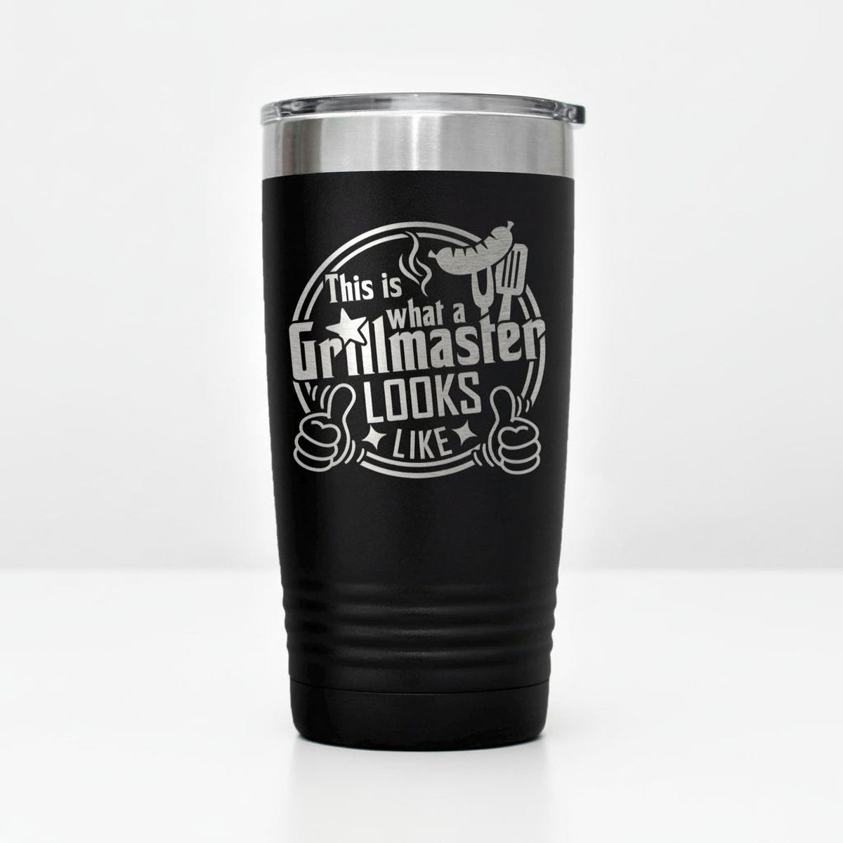 A black tumbler with a silver lid, featuring a humorous text and graphic design that reads "This is what a Grillmaster looks like" and includes a thumbs up symbol.