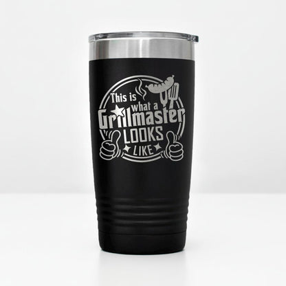 A black tumbler with a silver lid, featuring a humorous text and graphic design that reads "This is what a Grillmaster looks like" and includes a thumbs up symbol.