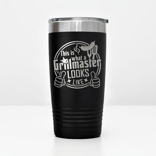 A black tumbler with a silver lid, featuring a humorous text and graphic design that reads "This is what a Grillmaster looks like" and includes a thumbs up symbol.