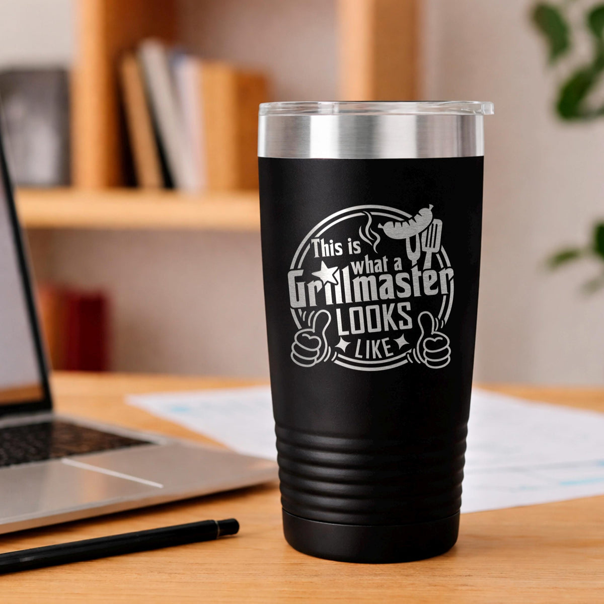 A black tumbler with a printed design on it, sitting on a wooden desk next to a laptop and a pen.