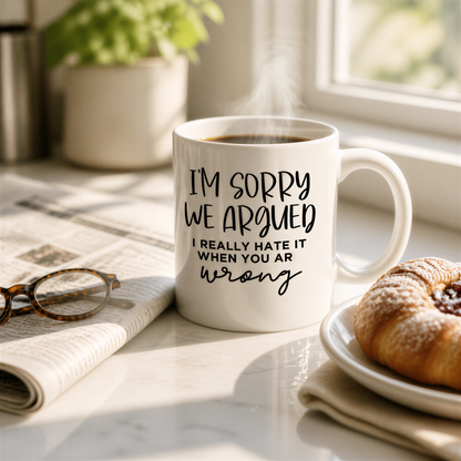 A white coffee mug with a humorous text printed on it, placed on a table next to a newspaper, a pastry, and a pair of glasses.