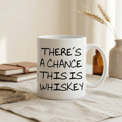 A white mug with black text that reads "THERE'S A CHANCE THIS IS WHISKEY" is placed on a table, with a stack of books and a vase of wheat stalks in the background.