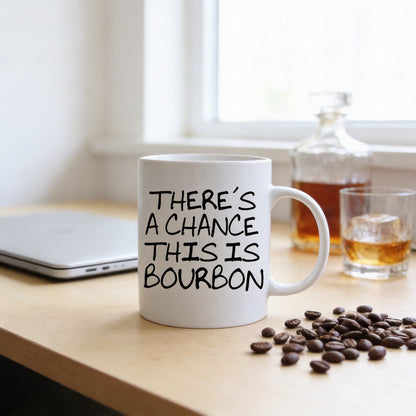 a white mug with the text "THERE'S A CHANCE THIS IS BOURBON" printed on it, placed on a wooden table next to a laptop and coffee beans.