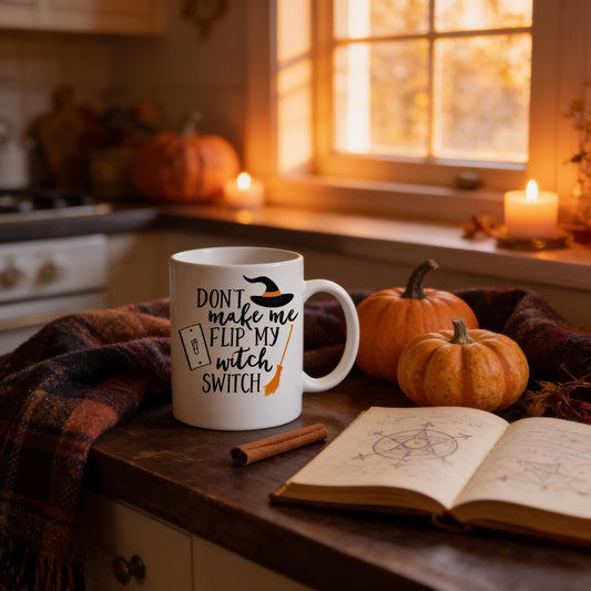 A mug with a humorous text that says "DON'T make me flip my witch switch" is placed on a kitchen counter, surrounded by pumpkins and a candle. An open book and a cinnamon stick are also present on the counter.