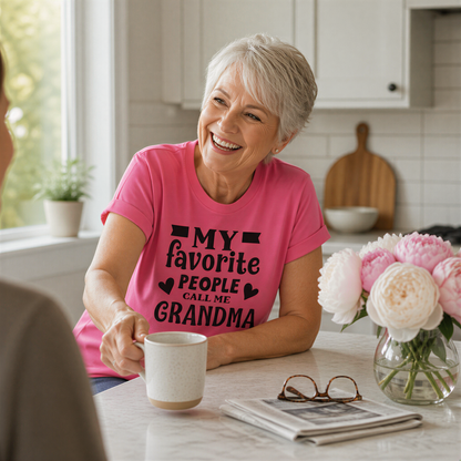 A smiling older woman wearing a pink t-shirt with text, sitting at a kitchen counter with a coffee mug, flowers, and glasses.