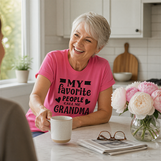 A smiling older woman wearing a pink t-shirt with text, sitting at a kitchen counter with a coffee mug, flowers, and glasses.