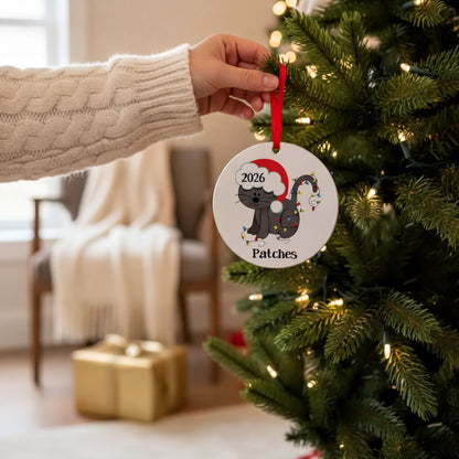 A hand is holding a Christmas ornament with a cat wearing a Santa hat and the number "2026" on it, hanging from a decorated Christmas tree.